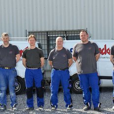 Eine Gruppe von sieben Männern in blauen Arbeitsanzügen steht auf einem Kiesplatz vor einem Firmenfahrzeug.