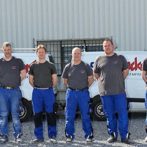 Eine Gruppe von sieben Männern in blauen Arbeitsanzügen steht auf einem Kiesplatz vor einem Firmenfahrzeug.