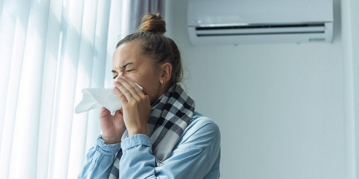 Im Vordergrund ist eine Frau in einem Raum, die Ihre Nase mit einem Taschentuch putzt. Im Hintergrund sieht man eine Klimaanlage an der Wand.