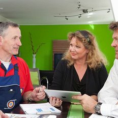 Der Handwerker, gekleidet in Arbeitskleidung, zeigt dem Paar auf einem Tablet wichtige Informationen während eines Beratungsgesprächs.