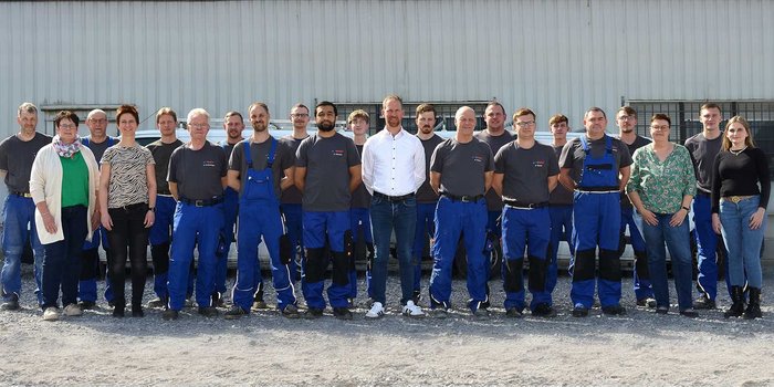 Mitarbeiter in blauen Overalls und T-Shirts posieren gemeinsam vor einem Firmengebäude, einige tragen Brillen.