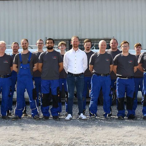 Mitarbeiter in blauen Overalls und T-Shirts posieren gemeinsam vor einem Firmengebäude, einige tragen Brillen.