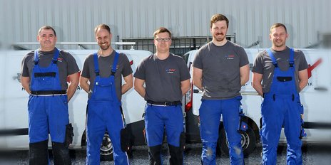 Fünf Männer in blauen Overalls stehen lächelnd vor einem Lieferwagen, bereit für ihre Arbeit im Freien.