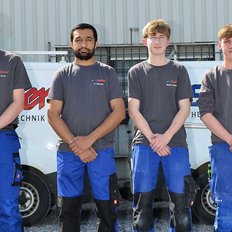 Vier Handwerker stehen stolz vor ihrem Firmenfahrzeug, gekleidet in graue T-Shirts und blaue Arbeitshosen, bereit für den Einsatz.