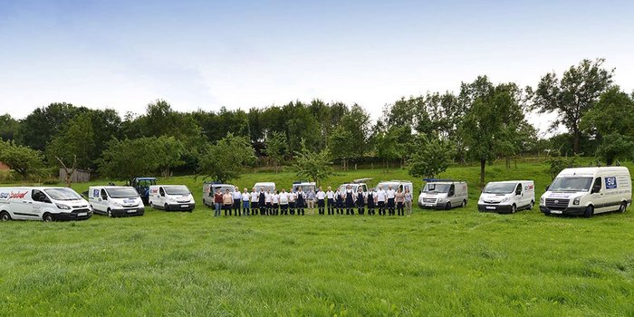Eine Gruppe von Mitarbeitern steht in einer Wiese, umgeben von mehreren weißen Lieferwagen und Bäumen im Hintergrund.