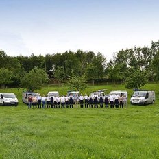 Eine Gruppe von Mitarbeitern steht in einer Wiese, umgeben von mehreren weißen Lieferwagen und Bäumen im Hintergrund.