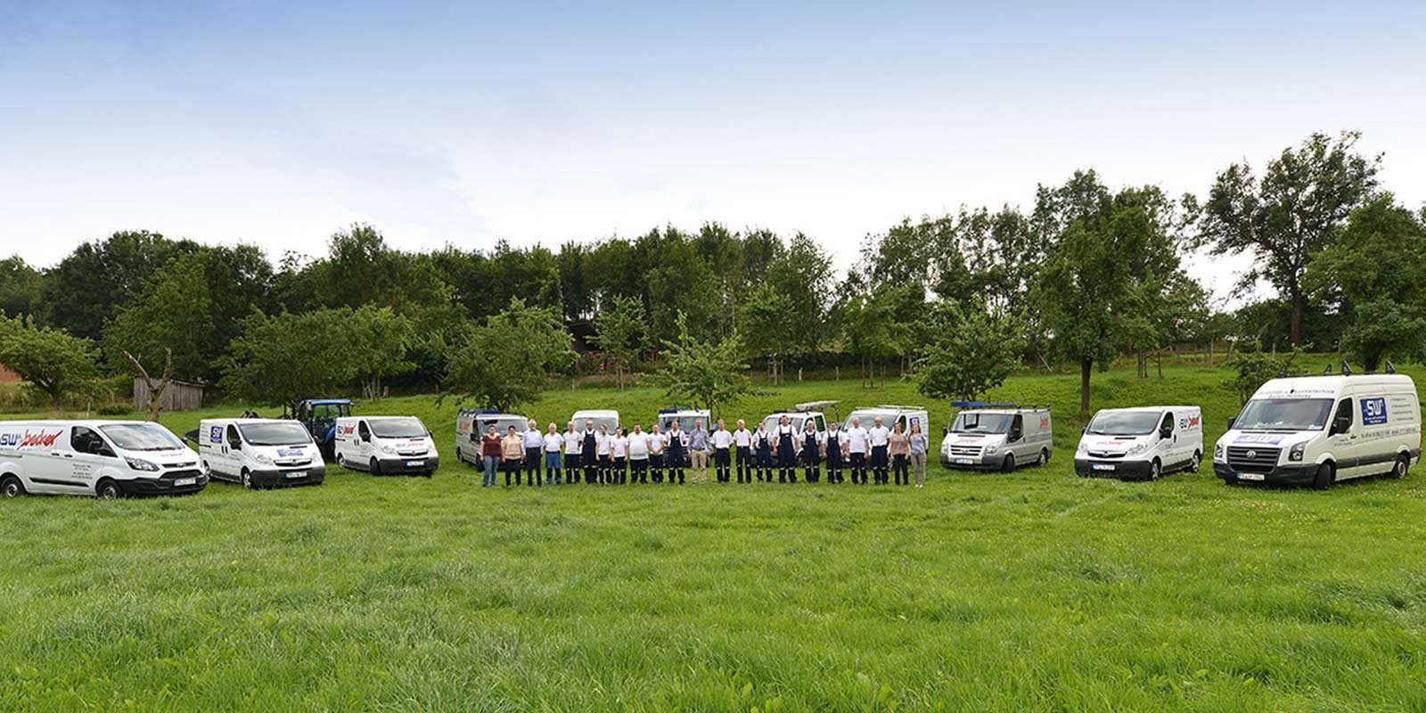 Eine Gruppe von Mitarbeitern steht in einer Wiese, umgeben von mehreren weißen Lieferwagen und Bäumen im Hintergrund.