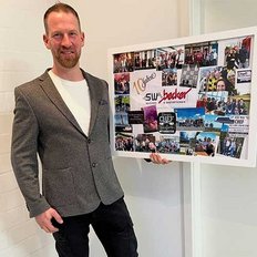 Ein Mann in einem grauen Blazer steht stolz vor einer Collage mit Fotos von Team-Events und besonderen Momenten.